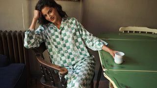 Model wears a green gingham check pyjama set at a table with a coffee cup on it. 