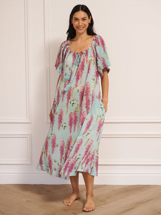 model wearing a duck egg blue cotton gauze maxi night dress with pink wisteria print, loose short sleeves, tassel tie neckline and relaxed fit, standing barefoot indoors.