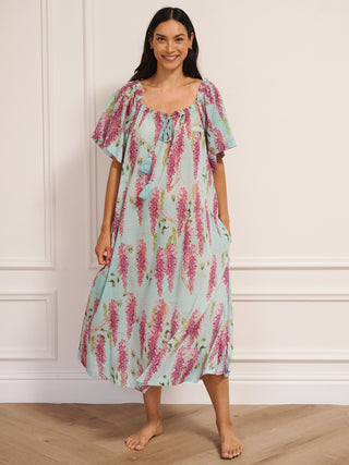 model wearing a duck egg blue cotton gauze maxi night dress with pink wisteria print, loose short sleeves, tassel tie neckline and relaxed fit, standing barefoot indoors.