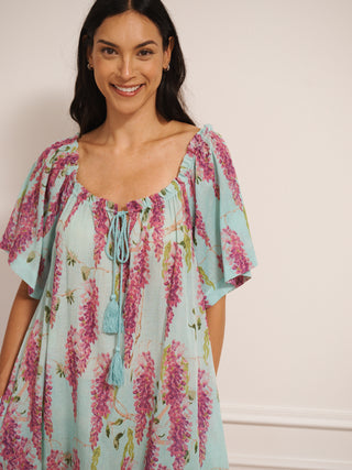 model wearing a duck egg blue cotton gauze maxi night dress with pink wisteria print, loose short sleeves, tassel tie neckline and relaxed fit, standing barefoot indoors.