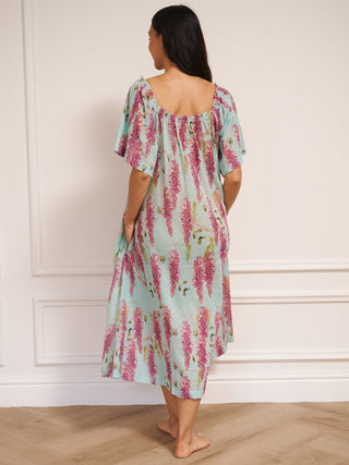 model wearing a duck egg blue cotton gauze maxi night dress with pink wisteria print, loose short sleeves, tassel tie neckline and relaxed fit, standing barefoot indoors.