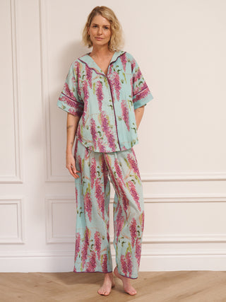 Model wearing a duck egg blue wisteria print pyjama set with a relaxed short-sleeve shirt and wide-leg trousers, standing barefoot in a bright, white room.