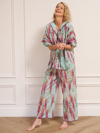 Model wearing a duck egg blue wisteria print pyjama set with a relaxed short-sleeve shirt and wide-leg trousers, standing barefoot in a bright, white room. model laughs
