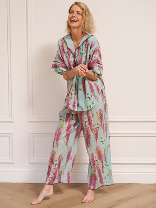 Woman wearing duck egg blue cotton gauze oversized pyjamas with pink wisteria floral print, short-sleeve button-through top and wide-leg trousers, standing barefoot indoors.