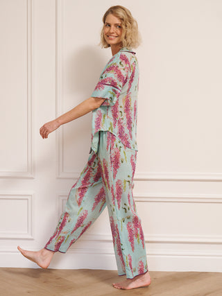 Model wearing a duck egg blue wisteria print pyjama set with a relaxed short-sleeve shirt and wide-leg trousers, standing barefoot in a bright, white room. side shot