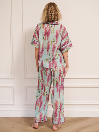 Model wearing a duck egg blue wisteria print pyjama set with a relaxed short-sleeve shirt and wide-leg trousers, standing barefoot in a bright, white room. back shot