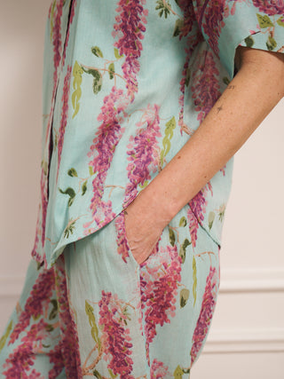 Model wearing a duck egg blue wisteria print pyjama set with a relaxed short-sleeve shirt and wide-leg trousers, standing barefoot in a bright, white room. detail shot
