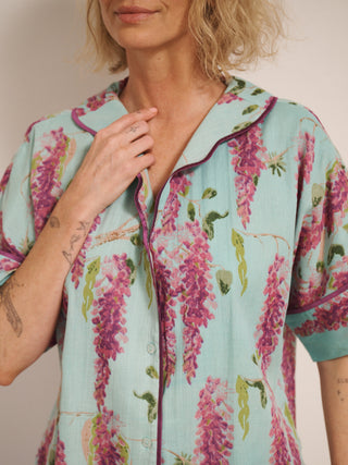 Model wearing a duck egg blue wisteria print pyjama set with a relaxed short-sleeve shirt and wide-leg trousers, standing barefoot in a bright, white room. detail shot