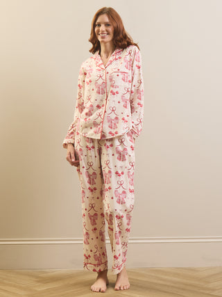 A woman modeling a two-piece pyjama set with a pink bow and ribbon pattern on a cream-colored fabric, standing against a beige wall and light wood floor.