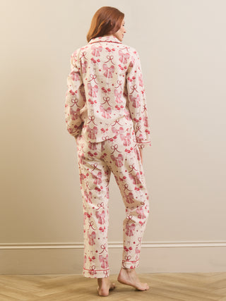 A woman stands barefoot indoors wearing a matching pyjama set with a pink bow and ribbon print on a light cream background, photographed from the back against a plain beige wall and wood floor.