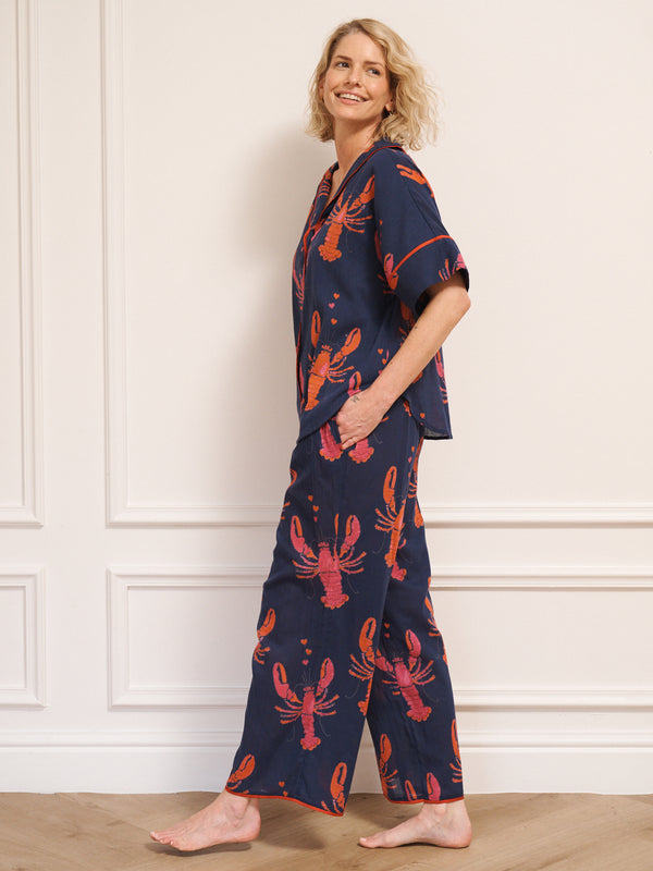 Model wearing women’s navy lobster print oversized pyjamas with short-sleeve button-up top and relaxed wide-leg trousers, photographed barefoot against a white studio wall.