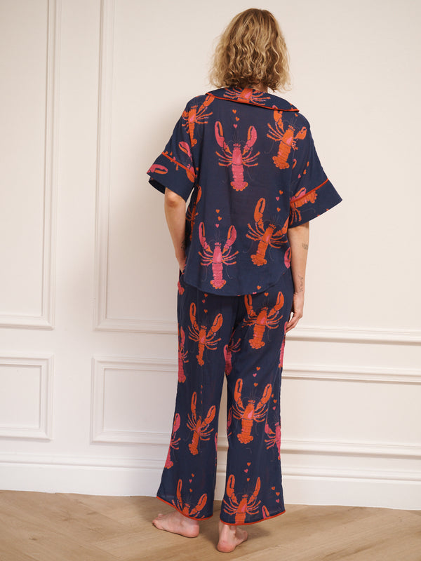 Model wearing women’s navy lobster print oversized pyjamas with short-sleeve button-up top and relaxed wide-leg trousers, photographed barefoot against a white studio wall. Back detail