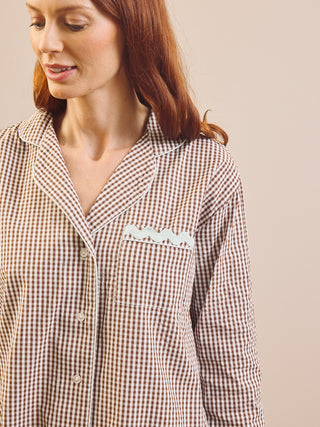 A woman wearing a brown and beige gingham pyjama nightshirt with a white embroidered wave detail on the chest pocket, set against a solid beige background.