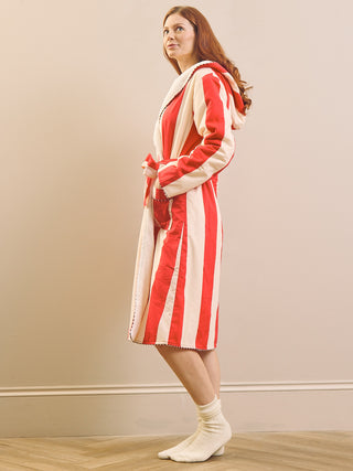 A woman wearing a red and cream striped hooded dressing gown bathrobe, standing in profile against a beige wall on a light wood floor.