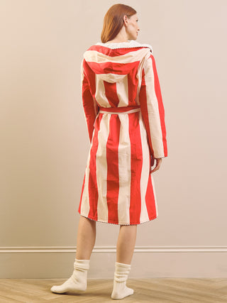 A model is shown from the back wearing a long-sleeved, belted dressing gown coat with bold red and cream vertical stripes, paired with cream socks, against a neutral beige wall and wooden floor.