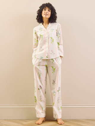 Woman wearing a light pink long payama set with floral pattern standing against a plain wall.
