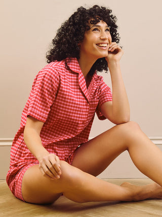 Woman in a red gingham short pyjamas set sitting on a wooden floor against a beige wall.