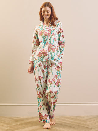 A woman modeling a two-piece pyjama set with a long-sleeve button-up top and matching pants, featuring a pink and green floral tulip pattern on a cream background, set against a neutral beige wall and light wood floor.