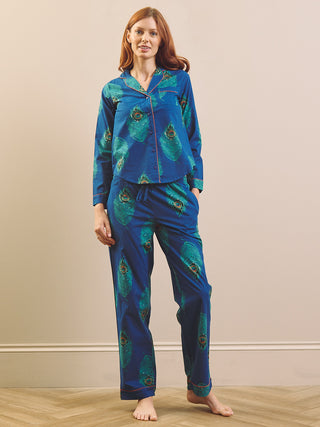 A woman modelling a matching two-piece pyjama set with a vibrant blue base and a green peacock feather print, standing against a neutral beige wall.