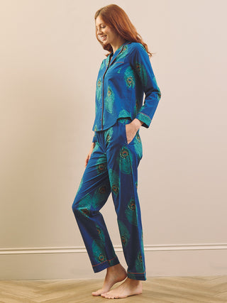 A woman modelling a two-piece pyjama set with a blue and teal peacock feather print, standing barefoot against a beige wall.