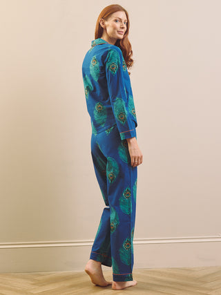A woman modelling a two-piece pyjama set with a blue and green peacock feather print, standing barefoot against a beige wall.