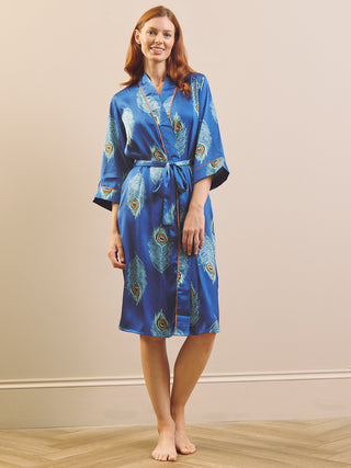 A woman stands barefoot in a studio wearing a blue satin dressing gown robe with a peacock feather print and a matching tie belt.