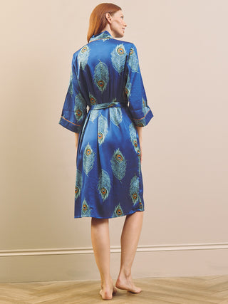 A woman stands barefoot in a studio wearing a blue satin dressing gown robe with a peacock feather print and a matching tie belt.