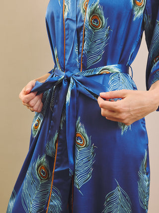 A close-up of a person wearing a blue satin dressing gown robe with a peacock feather print and a matching tie belt, adjusting the waist tie with both hands against a plain beige background.