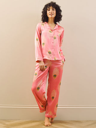 Woman wearing pink pyjama set with peacock feather patterns against a beige wall.