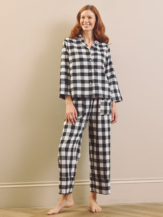 A woman modelling a two-piece pyjama set with a black and white gingham check pattern, standing against a beige wall and light wood floor.