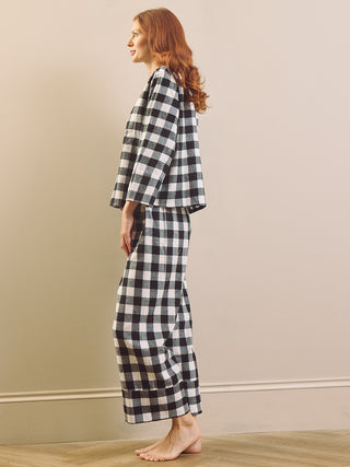 A woman with red hair stands in profile wearing a black and white gingham plaid boxy pyjama set, barefoot on a light wood floor against a beige wall.