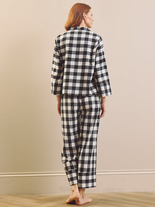 A woman stands barefoot against a beige wall, viewed from the back, wearing a matching black and white gingham check pyjama set with a long-sleeve top and cropped pants.