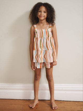 Young girl wearing a colorful striped dress standing against a plain wall.