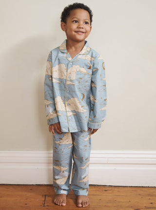 Child wearing blue cloudy sky print pyjamas standing on a wooden floor against a white wall.