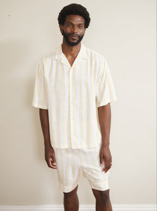 Model wearing a men’s buttermilk stripe linen-blend short pyjama set with a short-sleeve button-through shirt and matching shorts, featuring pale yellow and white vertical stripes, photographed barefoot against a neutral wall.