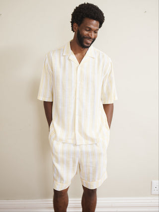 Model wearing a men’s buttermilk stripe linen-blend short pyjama set with a short-sleeve button-through shirt and matching shorts, featuring pale yellow and white vertical stripes, photographed barefoot against a neutral wall.