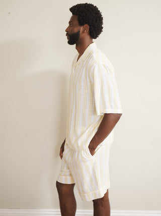 Model wearing a men’s buttermilk stripe linen-blend short pyjama set with a short-sleeve button-through shirt and matching shorts, featuring pale yellow and white vertical stripes, photographed barefoot against a neutral wall.