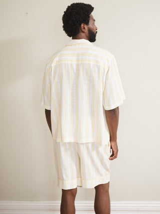Model wearing a men’s buttermilk stripe linen-blend short pyjama set with a short-sleeve button-through shirt and matching shorts, featuring pale yellow and white vertical stripes, photographed barefoot against a neutral wall.