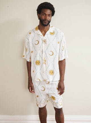 Model wearing a white men’s cotton short pyjama set with hand-drawn sun, moon and star print in yellow and blue, featuring a short-sleeve button-through shirt and matching shorts, photographed barefoot against a neutral wall.