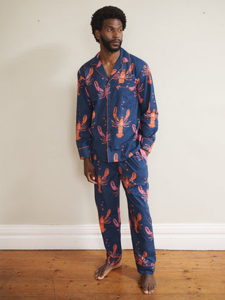 Model wearing a navy cotton lobster print pyjama set with coral lobsters and contrast piping, featuring a button-through long-sleeve top and matching trousers, photographed barefoot against a neutral wall.