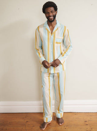 Model wearing a men’s cotton-linen long pyjama set with vertical aqua blue, mustard yellow and white stripes, featuring a button-through long-sleeve shirt with chest pocket and matching trousers.