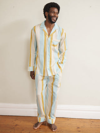 Model wearing a men’s cotton-linen long pyjama set with vertical aqua blue, mustard yellow and white stripes, featuring a button-through long-sleeve shirt with chest pocket and matching trousers.