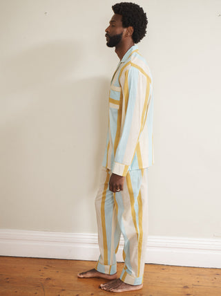Model wearing a men’s cotton-linen long pyjama set with vertical aqua blue, mustard yellow and white stripes, featuring a button-through long-sleeve shirt with chest pocket and matching trousers.