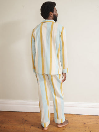 Model wearing a men’s cotton-linen long pyjama set with vertical aqua blue, mustard yellow and white stripes, featuring a button-through long-sleeve shirt with chest pocket and matching trousers. Back Detail. 