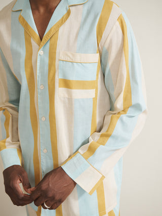 Model wearing a men’s cotton-linen long pyjama set with vertical aqua blue, mustard yellow and white stripes, featuring a button-through long-sleeve shirt with chest pocket and matching trousers.