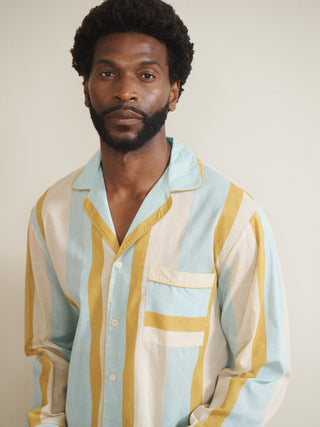Model wearing a men’s cotton-linen long pyjama set with vertical aqua blue, mustard yellow and white stripes, featuring a button-through long-sleeve shirt with chest pocket and matching trousers.