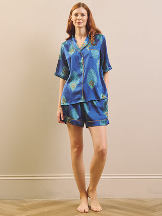 A woman stands barefoot indoors wearing a matching blue satin pyjama set with a teal and gold abstract print. The set includes a short-sleeve button-front top and coordinating shorts against a plain beige wall and light wood floor.