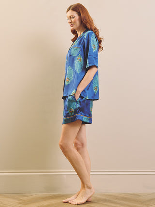 A woman stands barefoot in a room wearing a blue satin pyjama set with a peacock feather print. The set includes a short-sleeve button-up shirt and matching shorts, shown from a rear three-quarter angle against a beige wall and light wood floor.