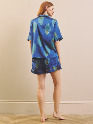 A woman stands barefoot in a room wearing a blue satin pyjama set with a peacock feather print. The set includes a short-sleeve button-up shirt and matching shorts, shown from a rear three-quarter angle against a beige wall and light wood floor.