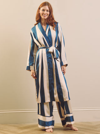 A woman modelling a long-sleeve satin dressing gown robe and matching pyjama pants with vertical blue and cream stripes, standing barefoot on a light wood floor against a beige wall.